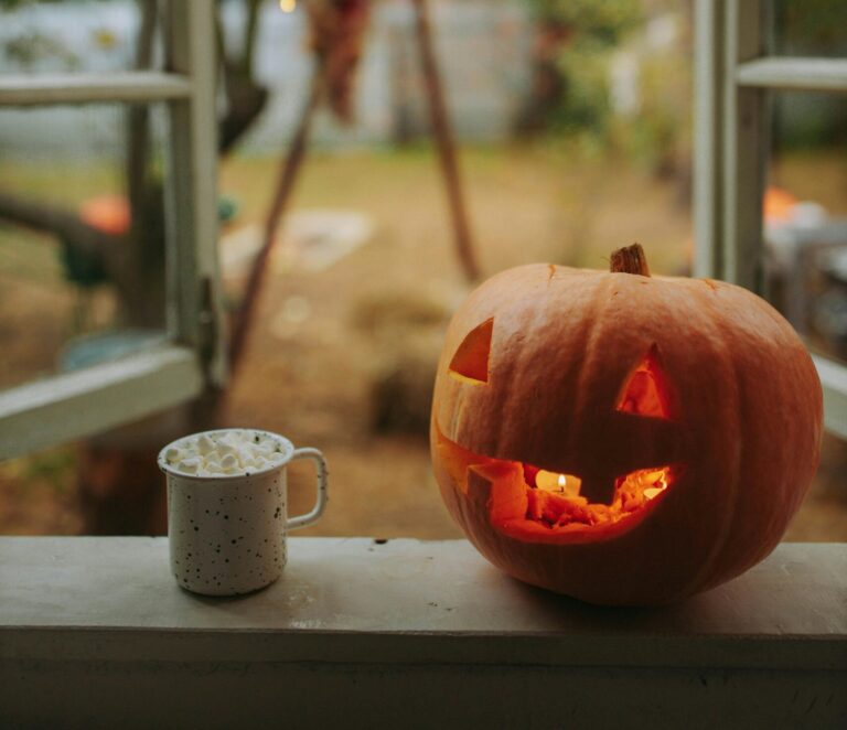 Halloween pumpkin and cozy mug on a window sill with string lights and fall scenery.